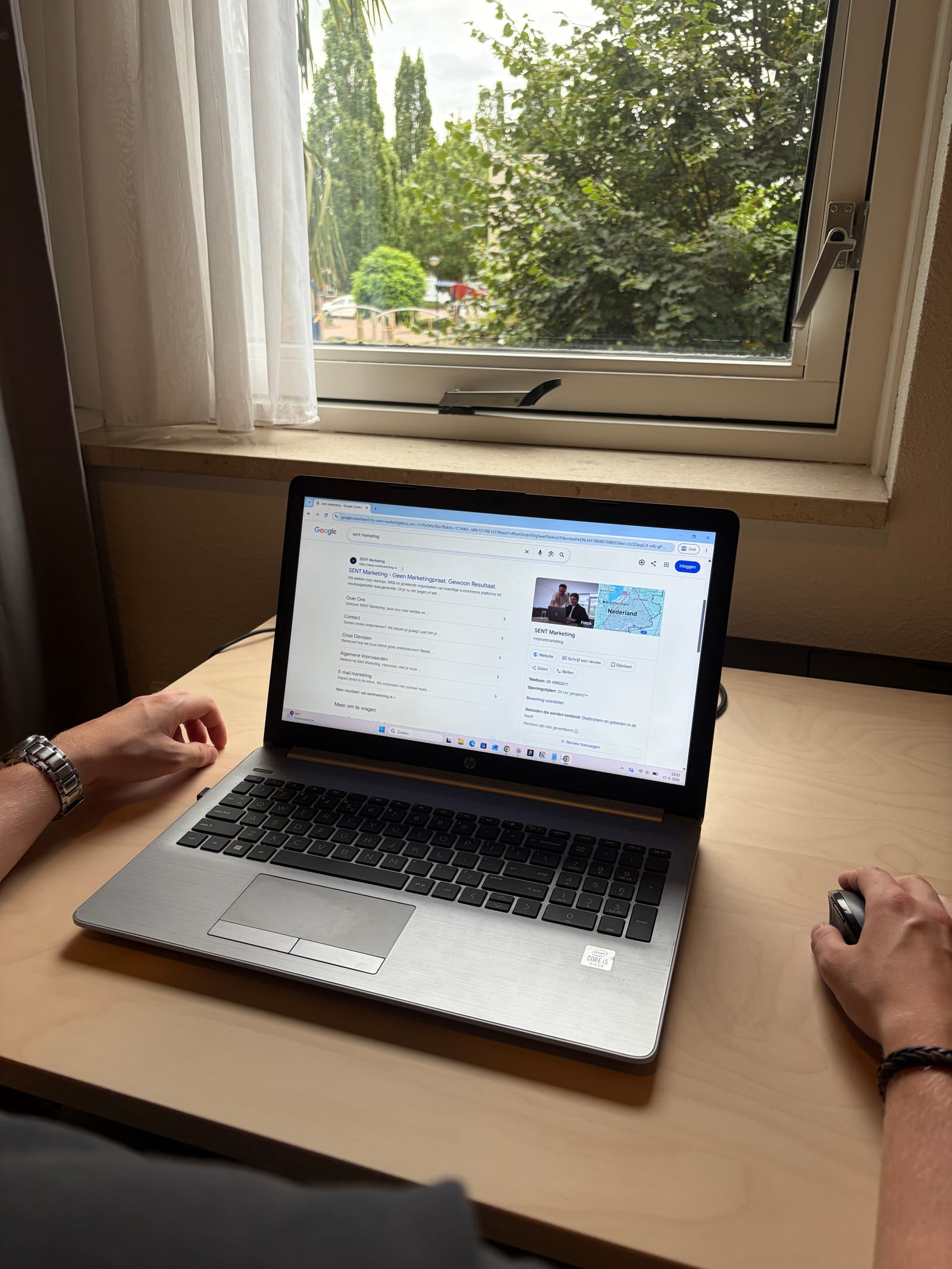 Laptop op een houten bureau bij een raam toont een Google-zoekresultaat voor SENT Marketing, met twee handen ernaast