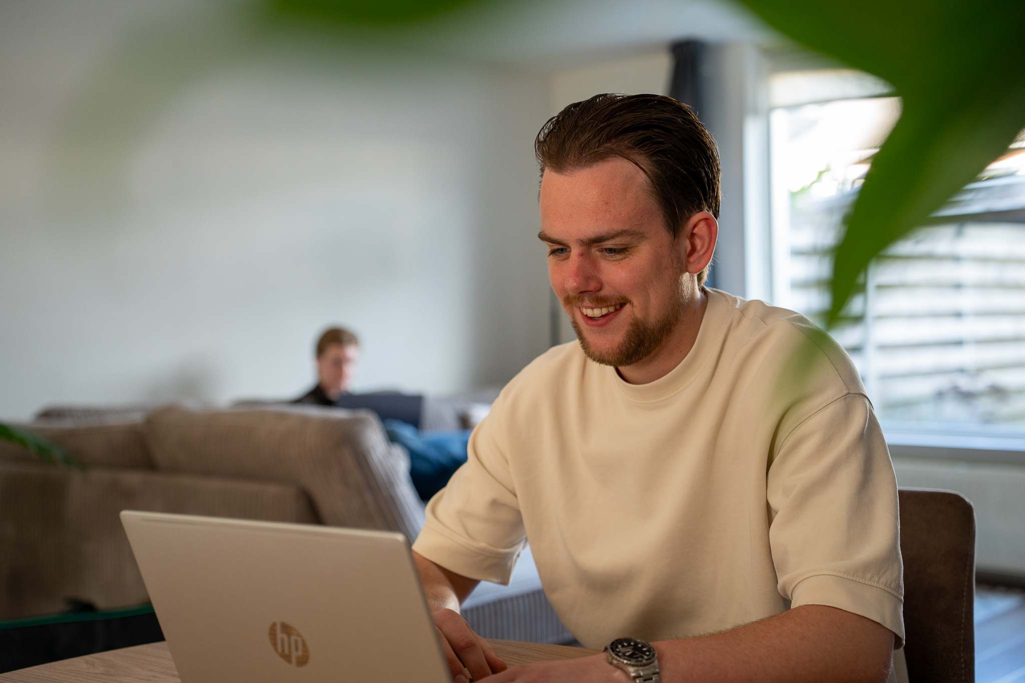 Glimlachende marketeer werkt op een laptop aan tafel, met op de achtergrond een collega op de bank