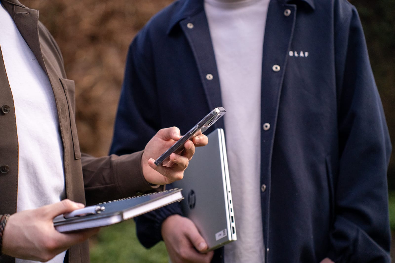Twee mensen buiten; de één kijkt op een smartphone met een notitieboek in de hand, de ander houdt een laptop vast
