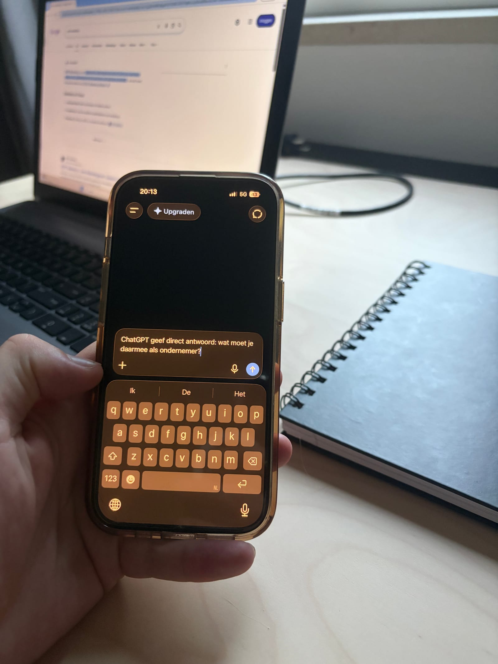 Hand houdt telefoon met ChatGPT-app vast voor een laptop op een bureau